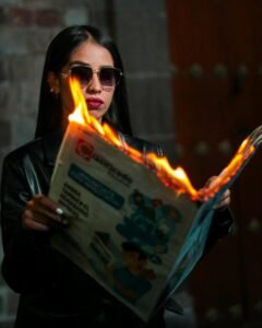 Fashionable woman in sunglasses reads burning newspaper indoors, blending style with danger.