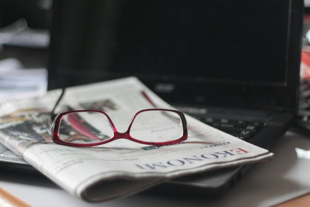 La Crecente Dominación de las Plataformas Digitales sobre los Diarios Tradicionales y su Influencia en la Opinión Pública Close-up of red eyeglasses on a newspaper with a laptop in the background, suggesting reading or work.
